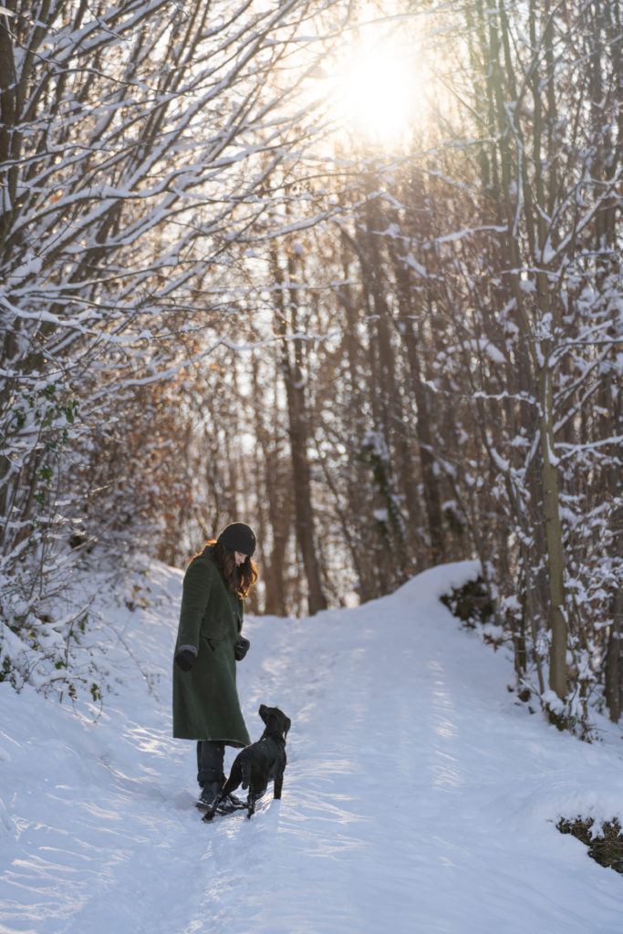 Eine Frau in einem grünen Mantel und einer Mütze geht mit ihrem schwarzen Hund auf einem verschneiten Waldweg, während die Wintersonne durch die Bäume scheint.