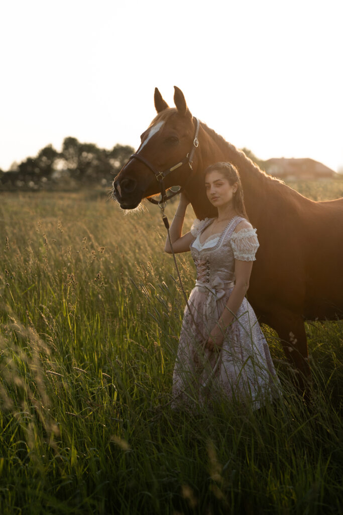 Eine junge Frau im Dirndl steht neben einem Pferd in einem Feld bei Sonnenuntergang, umgeben von hohem Gras.